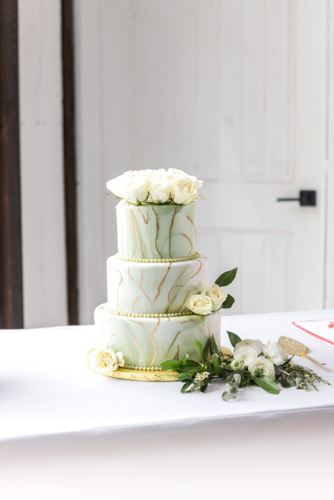 A beautiful sage and gold cake surrounded by flowers near bright open window at White Azalea Estate